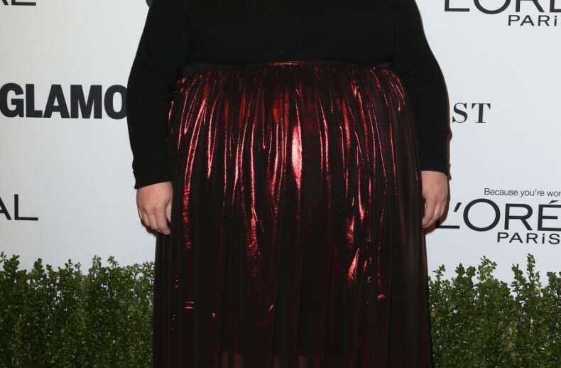 A person stands on a red carpet, embodying diversity in a black top and shiny red skirt. The background showcases logos for L'Oreal Paris and Glamour, as if part of their dynamic diversity programs.
