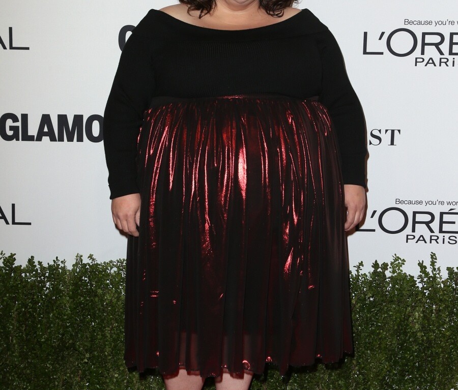 A person stands on a red carpet, embodying diversity in a black top and shiny red skirt. The background showcases logos for L'Oreal Paris and Glamour, as if part of their dynamic diversity programs.