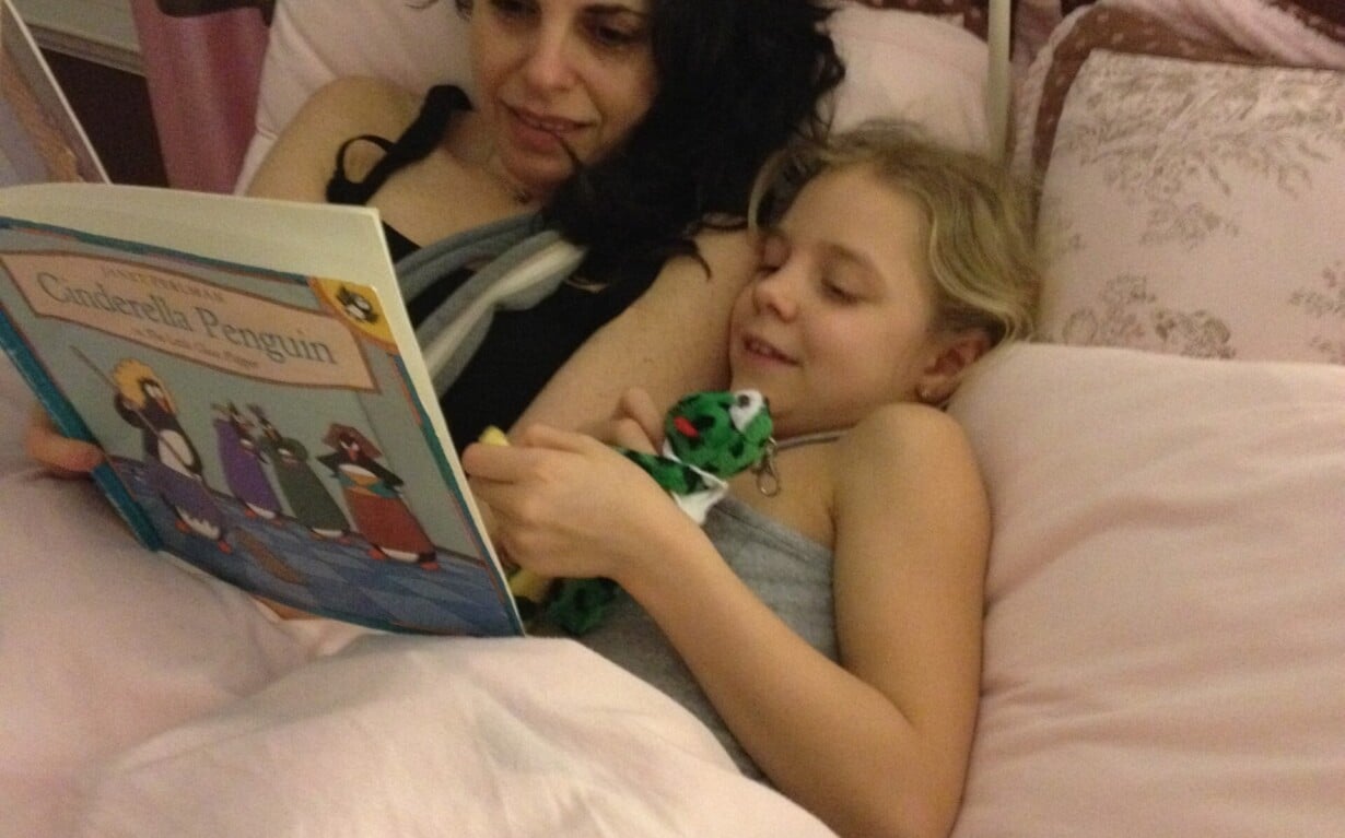 A woman and a child lie in bed under pink covers, engaging in social emotional learning activities. The woman reads a book titled "Costabella Penguin" to the child, who holds a small toy and smiles brightly, embracing quality time together.