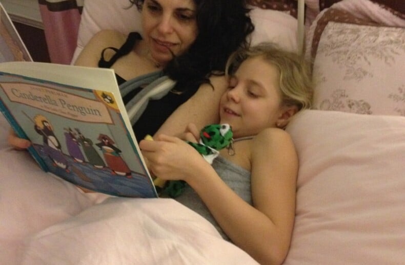 A woman and a child lie in bed under pink covers, engaging in social emotional learning activities. The woman reads a book titled "Costabella Penguin" to the child, who holds a small toy and smiles brightly, embracing quality time together.
