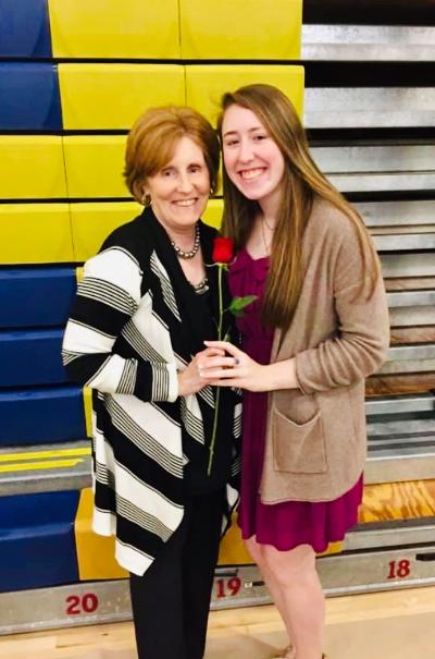 Two women stand smiling in front of a multicolored wall. The older woman, wearing a striped cardigan, holds a red rose. The younger woman beside her wears a pink dress and beige cardigan. Their joyful bond is reminiscent of the warmth fostered by SEL activities in nurturing relationships.