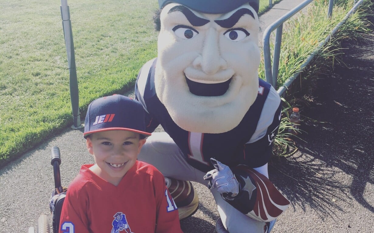 A young boy in a wheelchair, proudly wearing a football jersey and cap, poses with a team mascot on a sunny day. This heartwarming scene beside a grassy field highlights the impact of diversity programs in fostering inclusion and joy among all participants.