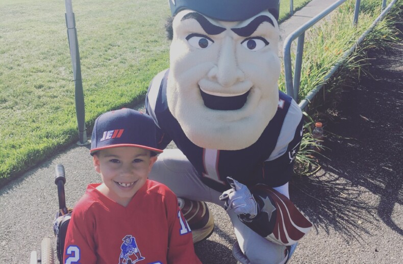 A young boy in a wheelchair, proudly wearing a football jersey and cap, poses with a team mascot on a sunny day. This heartwarming scene beside a grassy field highlights the impact of diversity programs in fostering inclusion and joy among all participants.