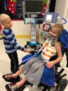 In a hospital room filled with medical devices, a young girl in a wheelchair, wearing a neck brace and clutching medical equipment, sits beside a young boy in a striped shirt who gently holds her hand. This tender moment highlights the essence of social emotional learning activities.