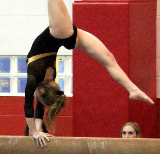 A gymnast performs a handstand on a balance beam in a gym, demonstrating amazing control and focus, much like the concentration often fostered in SEL activities. She wears a black and yellow leotard. Judges are seated at a table nearby, one seen holding a pen and paper.