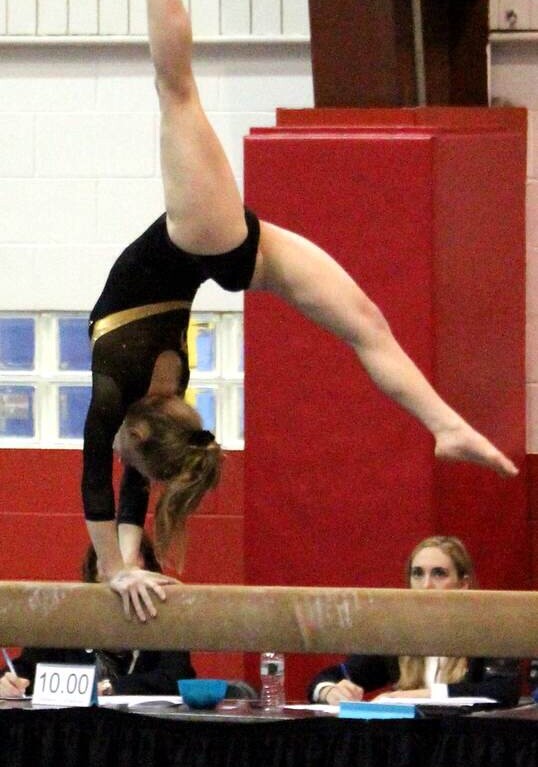 A gymnast performs a handstand on a balance beam in a gym, demonstrating amazing control and focus, much like the concentration often fostered in SEL activities. She wears a black and yellow leotard. Judges are seated at a table nearby, one seen holding a pen and paper.