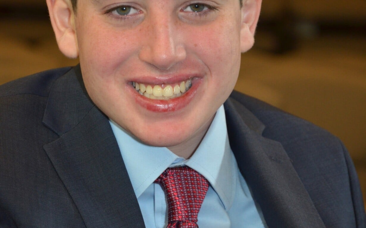 A young man in a blue suit and red tie smiles warmly at the camera, embodying the spirit of diversity programs. He has short brown hair and is seated in an indoor setting, where his engaging presence invites dialogue and learning.