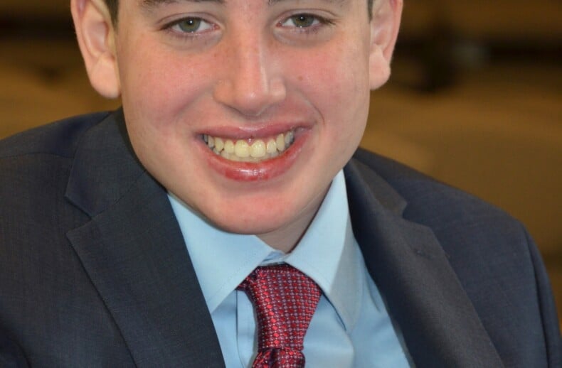 A young man in a blue suit and red tie smiles warmly at the camera, embodying the spirit of diversity programs. He has short brown hair and is seated in an indoor setting, where his engaging presence invites dialogue and learning.