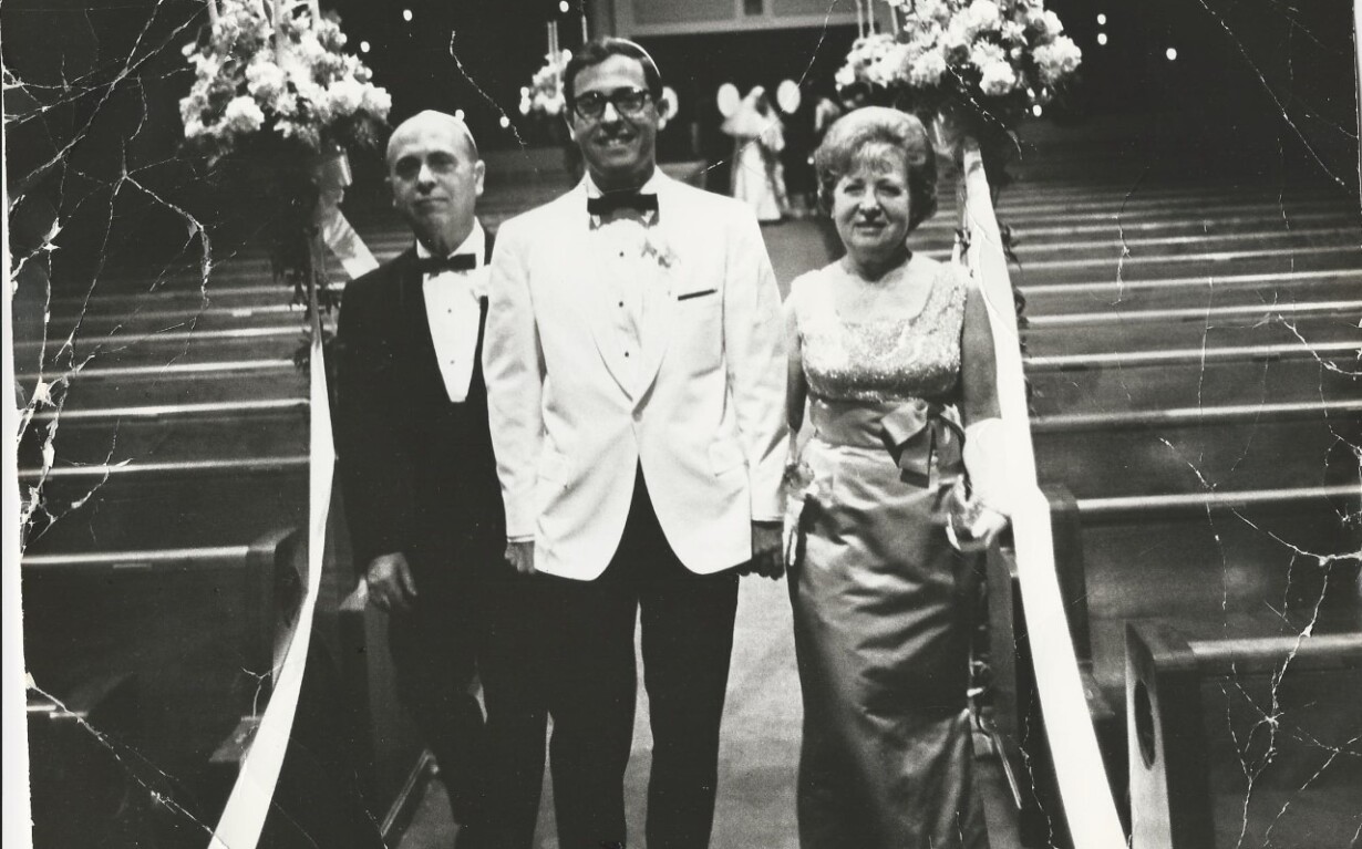 A groom in a white tuxedo stands between an older man and woman in formal attire, embodying the essence of social emotional learning activities through the grace of family bonds. At the church aisle entrance, flower arrangements adorn their path, symbolizing unity and love.