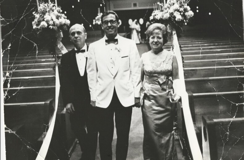 A groom in a white tuxedo stands between an older man and woman in formal attire, embodying the essence of social emotional learning activities through the grace of family bonds. At the church aisle entrance, flower arrangements adorn their path, symbolizing unity and love.