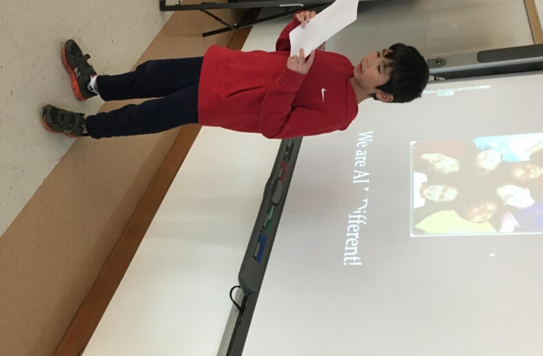 A child in a red sweater stands in front of a presentation screen, reading from a sheet of paper as part of an SEL activity. The screen shows a slide with a group photo and the text "We are all different!" The setting appears to be a classroom engaged in social emotional learning.
