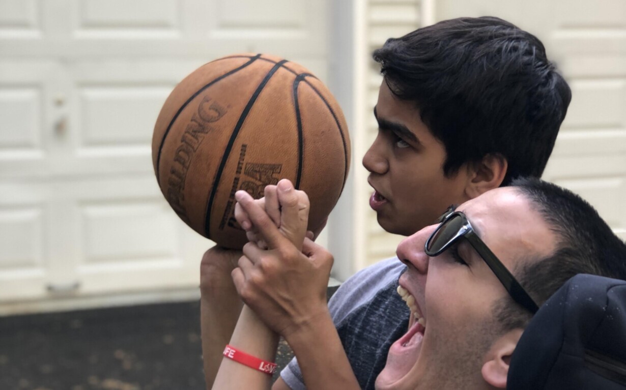 Two young men are outside in front of a garage. One is in a wheelchair, helping the other aim a basketball, illustrating the essence of social emotional learning activities. Both appear focused and engaged, highlighting simple yet profound lessons in teamwork and empathy.