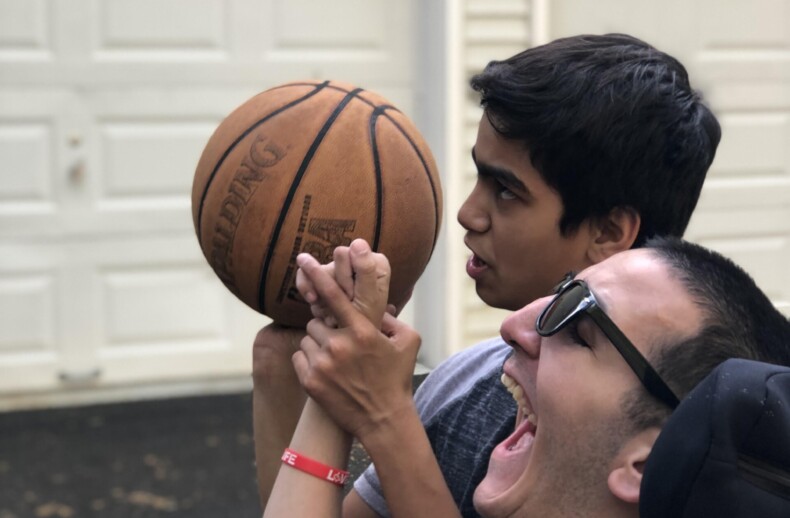 Two young men are outside in front of a garage. One is in a wheelchair, helping the other aim a basketball, illustrating the essence of social emotional learning activities. Both appear focused and engaged, highlighting simple yet profound lessons in teamwork and empathy.