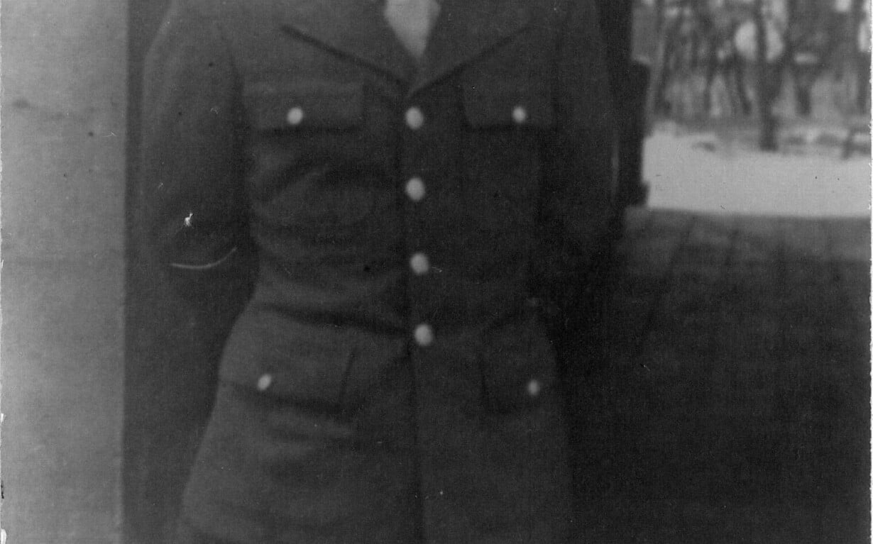 A man wearing a military uniform with a cap stands smiling in front of an arched doorway. His hands are behind his back, evoking the warmth of social-emotional learning activities. The blurred backdrop reveals trees and a path, captured elegantly in black and white.