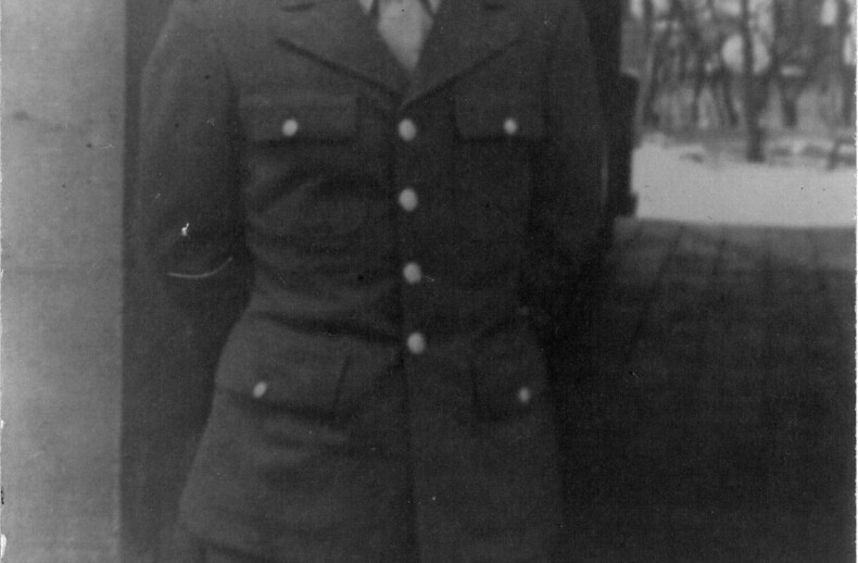 A man wearing a military uniform with a cap stands smiling in front of an arched doorway. His hands are behind his back, evoking the warmth of social-emotional learning activities. The blurred backdrop reveals trees and a path, captured elegantly in black and white.