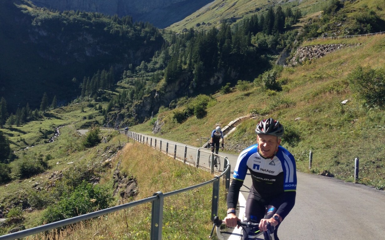 Cyclists ride uphill on a paved road in a mountainous landscape, an inspiring scene akin to social emotional learning activities. The lush green hills, steep hillside, and snow-capped mountain under the clear blue sky mirror the resilience and determination of the lead cyclist in blue and white.