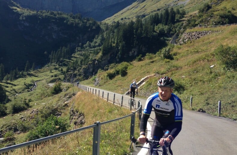 Cyclists ride uphill on a paved road in a mountainous landscape, an inspiring scene akin to social emotional learning activities. The lush green hills, steep hillside, and snow-capped mountain under the clear blue sky mirror the resilience and determination of the lead cyclist in blue and white.