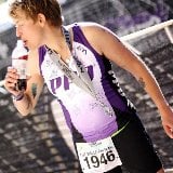 A person in a purple athletic outfit drinks from a bottle, proudly displaying their race number "1946" and medals around their neck, near a black netted barrier at the finish area of an event. This scene highlights the spirit of diversity programs that celebrate individual achievements.