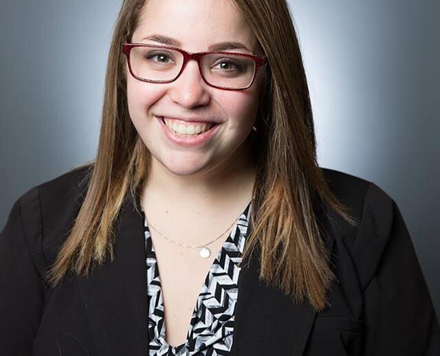 A woman with long, straight hair wearing red glasses smiles at the camera, embodying warmth and approachability. Dressed in a black blazer over a patterned top, she stands against a gray background, ready to engage in social emotional learning activities.