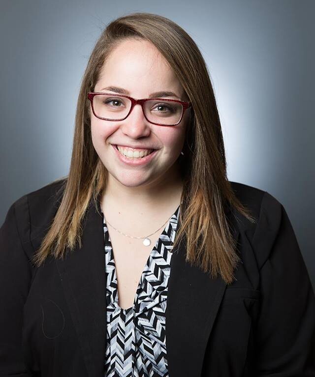 A woman with long, straight hair wearing red glasses smiles at the camera, embodying warmth and approachability. Dressed in a black blazer over a patterned top, she stands against a gray background, ready to engage in social emotional learning activities.