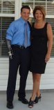A man in a blue shirt with a prosthetic arm and a tie stands next to a woman in a black dress, embodying the spirit of diversity programs. They pose joyfully in front of a building with white siding and windows, both smiling warmly.