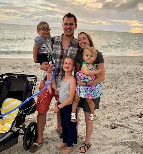 A family of five beams with joy at the beach during sunset. The man holds a young boy in his right arm, while a young girl stands in front of him. The woman cradles a toddler, and a stroller rests nearby on the sand—a perfect scene reflecting the values taught in SEL curriculum.