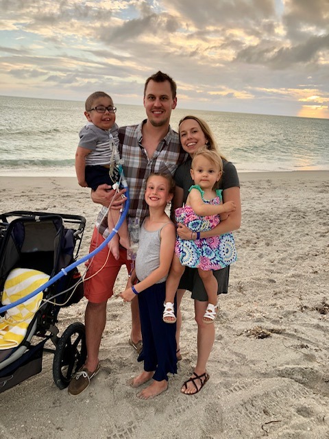 A family of five beams with joy at the beach during sunset. The man holds a young boy in his right arm, while a young girl stands in front of him. The woman cradles a toddler, and a stroller rests nearby on the sand—a perfect scene reflecting the values taught in SEL curriculum.
