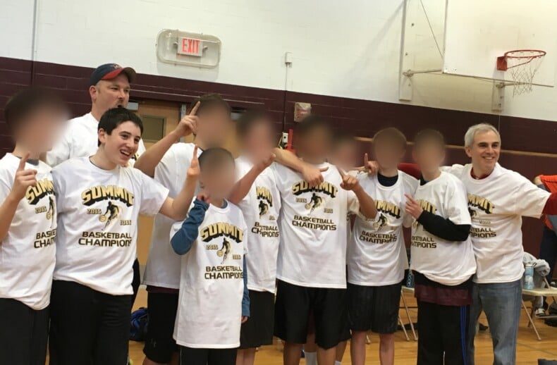A group of boys and two adults wearing matching "Basketball Champions" t-shirts pose in a gymnasium, celebrating their victory through teamwork and the positive impact of their social emotional learning curriculum. They stand in line, smiling with victory signs, as a basketball hoop looms behind them.