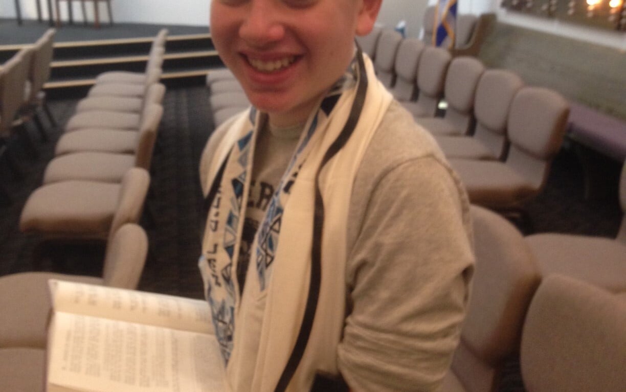 A smiling boy wearing a kippah, tallit, and tefillin holds an open prayer book in a synagogue, embodying values often highlighted in social emotional learning activities. Rows of empty chairs and an Israeli flag are in the background.