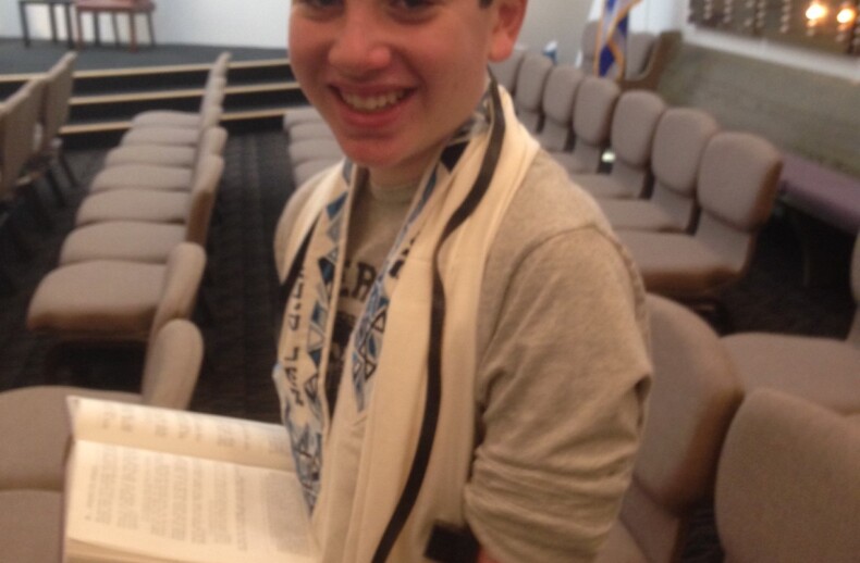 A smiling boy wearing a kippah, tallit, and tefillin holds an open prayer book in a synagogue, embodying values often highlighted in social emotional learning activities. Rows of empty chairs and an Israeli flag are in the background.