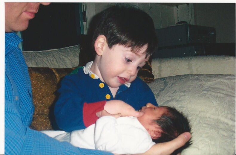 A young boy with dark hair and a blue shirt gently holds a newborn, wrapped in a white blanket, in his father's arms. As part of his school's SEL curriculum, he looks down lovingly at the baby, learning empathy and connection on their cozy couch.