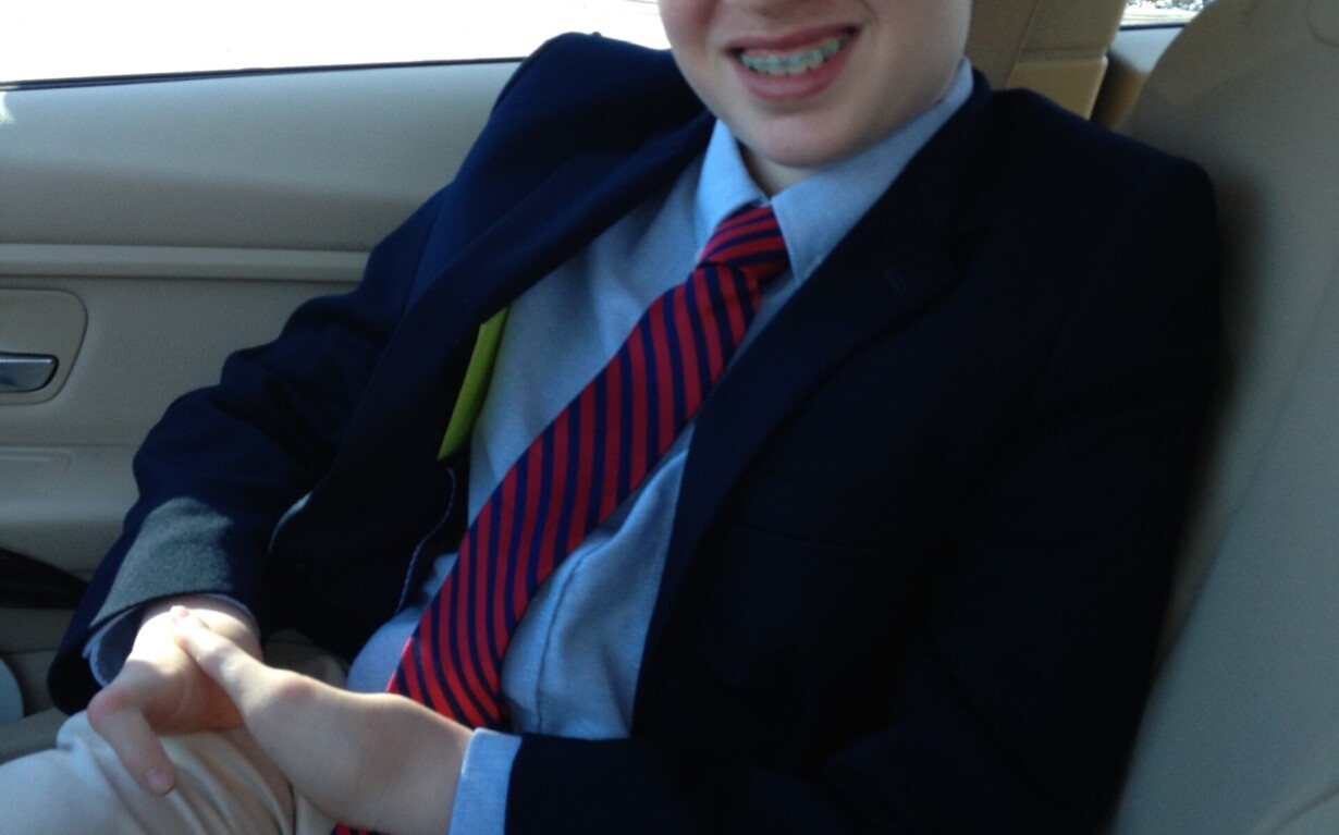 A young boy in a suit, blue shirt, and red-striped tie sits in the passenger seat of a car, smiling as he contemplates his day filled with engaging SEL activities. With hands resting on his lap, he looks forward to sharing what he's learned as trees blur by outside the window.