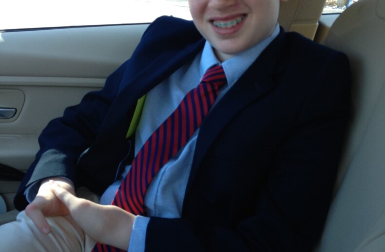 A young boy in a suit, blue shirt, and red-striped tie sits in the passenger seat of a car, smiling as he contemplates his day filled with engaging SEL activities. With hands resting on his lap, he looks forward to sharing what he's learned as trees blur by outside the window.