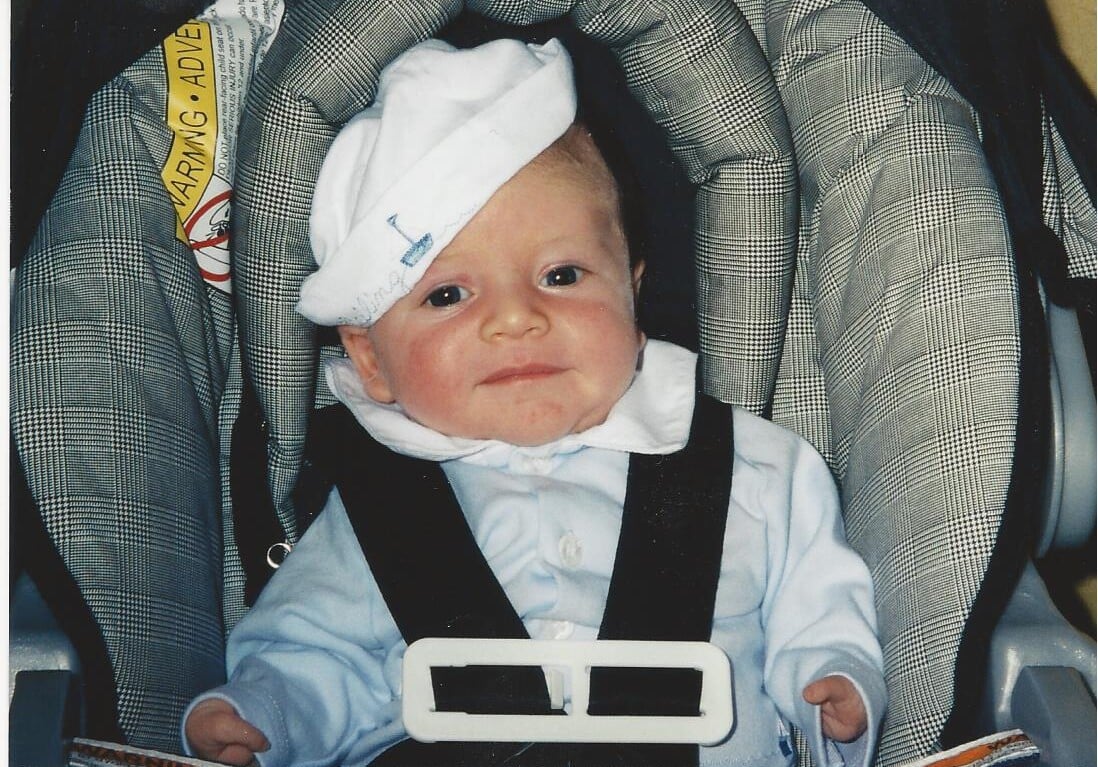 A baby in a car seat, wearing a white hat slightly tilted to the side and a blue outfit, is curiously gazing at the camera. The car seat features a gray and white checkered pattern, reminiscent of the playful setups during get-to-know-you activities for students, where every detail counts.