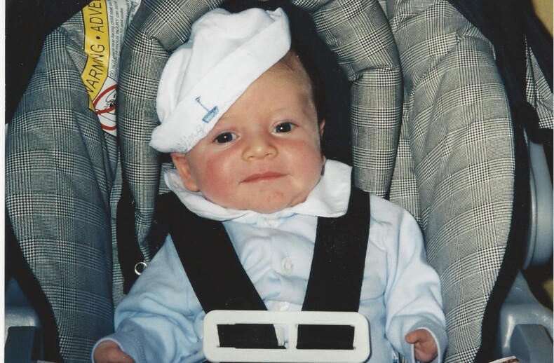 A baby in a car seat, wearing a white hat slightly tilted to the side and a blue outfit, is curiously gazing at the camera. The car seat features a gray and white checkered pattern, reminiscent of the playful setups during get-to-know-you activities for students, where every detail counts.