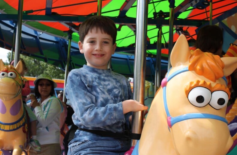 A boy with brown hair rides a colorful carousel horse, smiling at the camera. The vibrant, multicolored canopy mirrors the joy of SEL activities that encourage emotional growth. Other people are visible in the background, observing and sharing in the joyful experience.