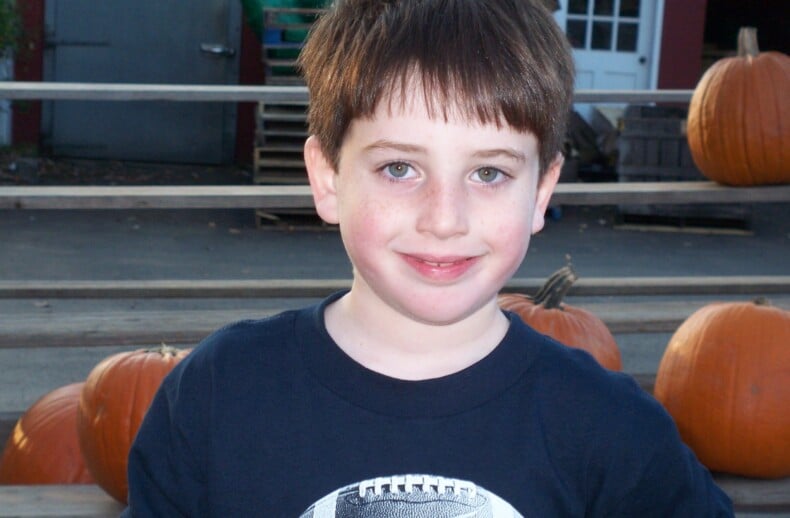 A young boy with brown hair smiles at the camera, embodying the joy found in social emotional learning activities. He's wearing a navy blue shirt with a football graphic. Behind him are several pumpkins and a wooden railing, while a red building and door add charm to the scene.
