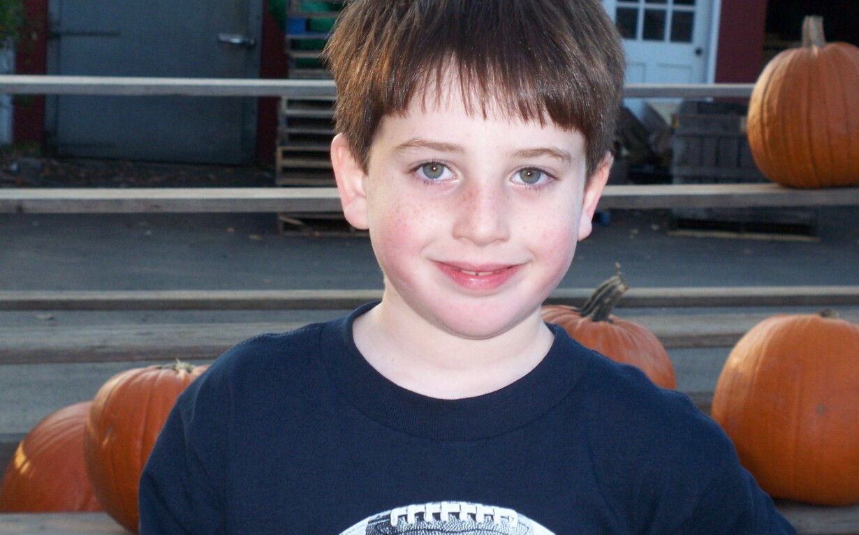 A young boy with brown hair and a blue shirt smiles while standing outdoors, his expression reflecting the joy of SEL activities. Behind him are several large orange pumpkins and a wooden railing, with a building featuring a red exterior and white door visible in the background.