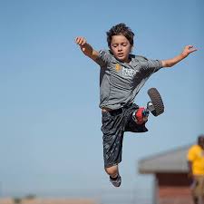 A young boy in athletic clothing is mid-air, jumping energetically against a clear blue sky, embodying the confidence and determination often nurtured through social emotional learning curriculum. He wears a gray shirt and black shorts, with one arm raised and a determined expression on his face.