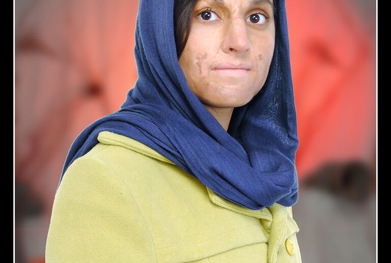 A woman wearing a blue headscarf and a mustard-yellow coat stands against a softly blurred background with red and white tones, embodying the spirit of diversity programs. She looks directly at the camera with a neutral expression.