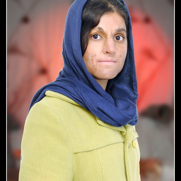 A woman wearing a blue headscarf and a mustard-yellow coat stands against a softly blurred background with red and white tones, embodying the spirit of diversity programs. She looks directly at the camera with a neutral expression.