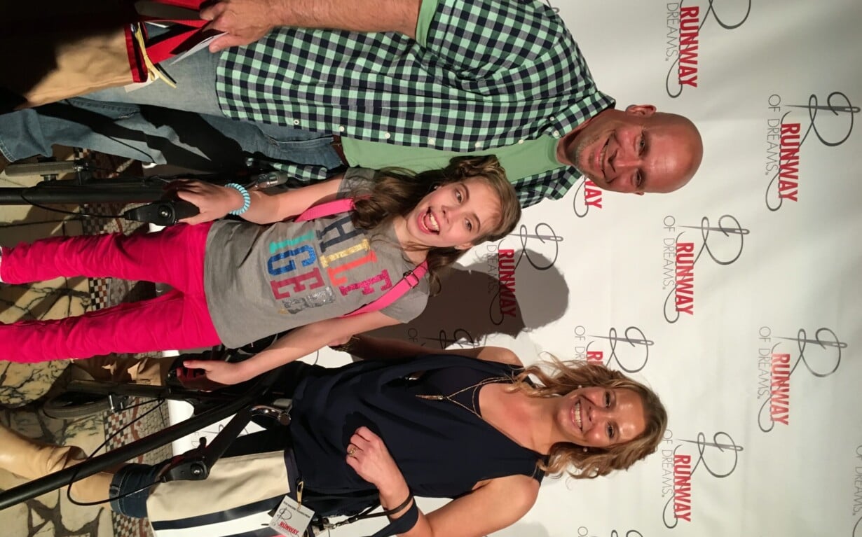 A smiling group of three people poses in front of a "Runway of Dreams" backdrop, embodying the spirit of diversity programs. The man sports a green checkered shirt, the girl in the middle dazzles in a colorful outfit, and the woman wears a chic dark sleeveless top.