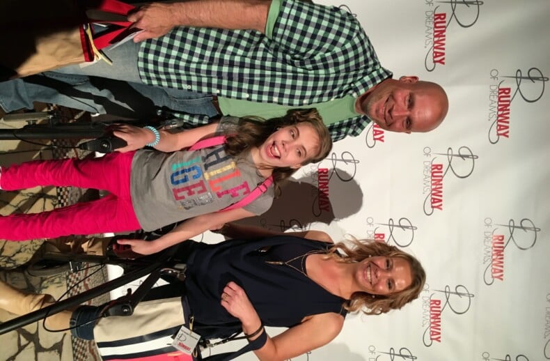 A smiling group of three people poses in front of a "Runway of Dreams" backdrop, embodying the spirit of diversity programs. The man sports a green checkered shirt, the girl in the middle dazzles in a colorful outfit, and the woman wears a chic dark sleeveless top.
