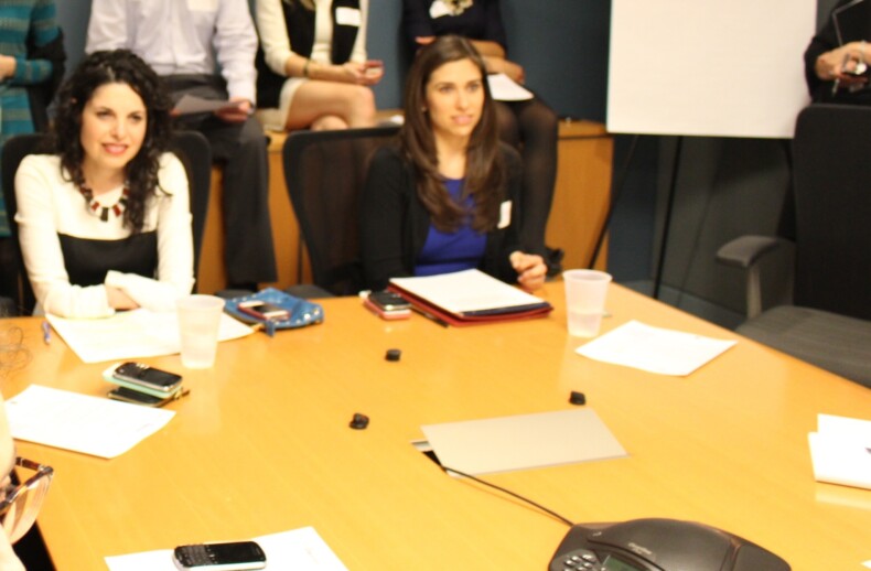 A group of people in business attire are seated around a conference table, engaged in what seems like diversity programs discussions. Two women are in focus, exchanging ideas over documents and phones. Others stand in the background, all set against a room filled with office furniture and a presentation board.