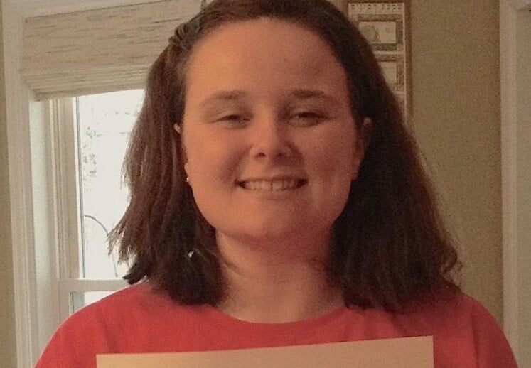 A person with shoulder-length hair smiles while holding a certificate, standing indoors with a window in the background. Their achievement highlights success in social emotional learning activities, showcasing commitment to fostering personal growth and understanding.