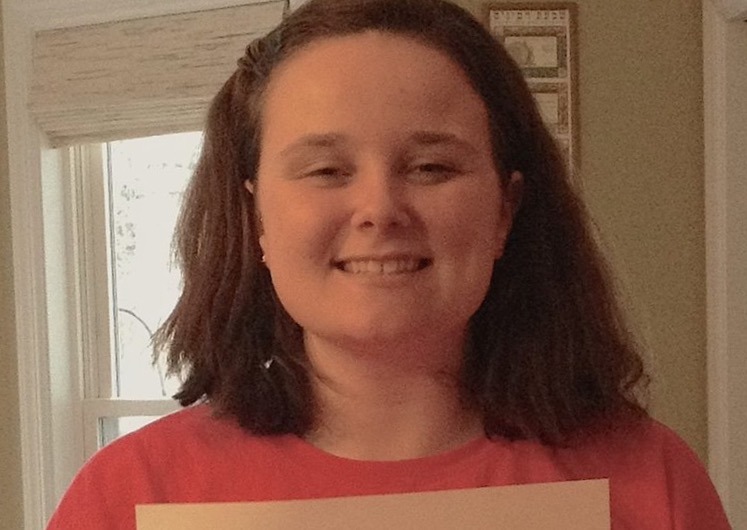 A person with shoulder-length hair smiles while holding a certificate, standing indoors with a window in the background. Their achievement highlights success in social emotional learning activities, showcasing commitment to fostering personal growth and understanding.