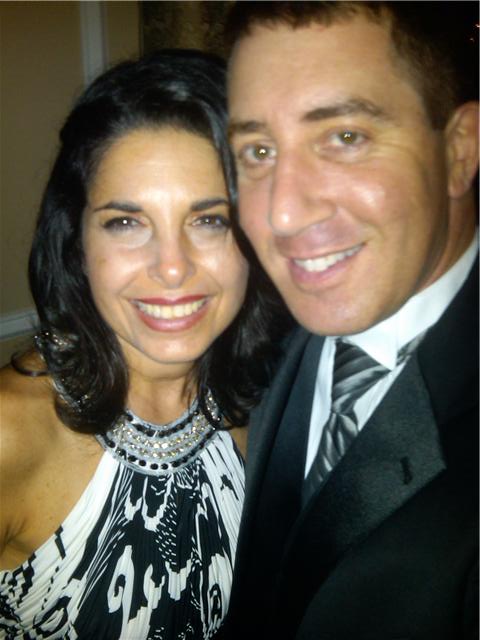 A woman and a man are smiling at the camera, exuding confidence. The woman, with long dark hair, dons a patterned dress with a jeweled neckline. The man sports a black suit and striped tie. They seem engaged in an event focused on SEL activities, promoting social emotional learning through connection.