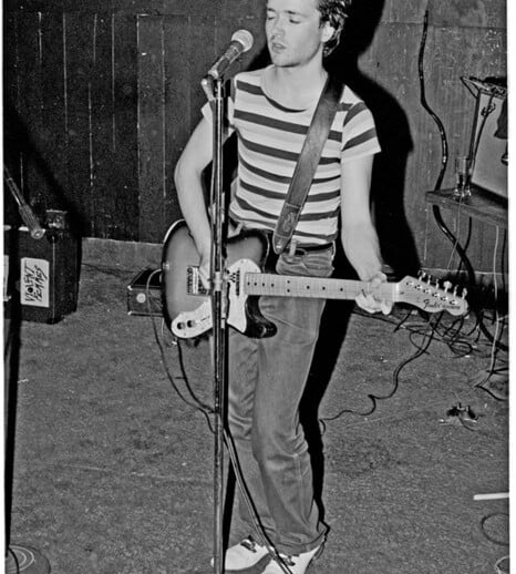 On stage, a person in a striped shirt and jeans plays an electric guitar and sings into the microphone. The dark, wooden background accentuates their performance, seamlessly blending creativity with expressions akin to social emotional learning activities.