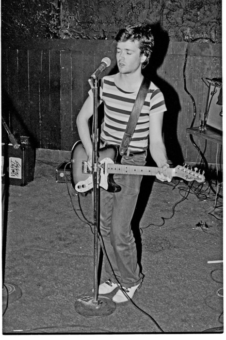 On stage, a person in a striped shirt and jeans plays an electric guitar and sings into the microphone. The dark, wooden background accentuates their performance, seamlessly blending creativity with expressions akin to social emotional learning activities.