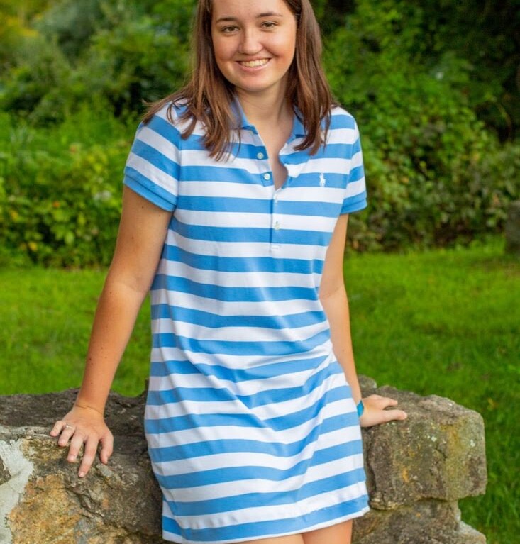 A woman in a blue and white striped dress smiles gracefully while leaning against a stone wall, embodying the serenity often fostered by a robust social emotional learning curriculum. Behind her, lush green foliage adds to the scene's peaceful ambiance.